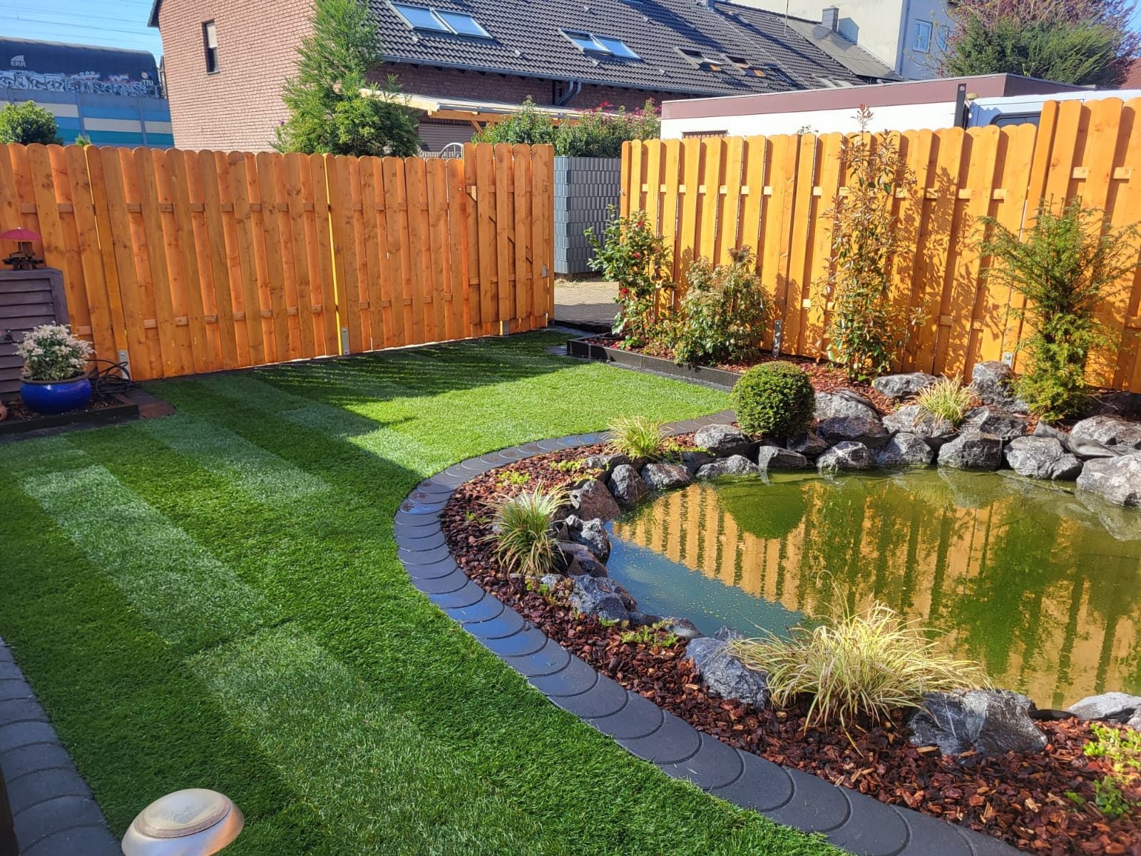 Garten mit Teich, Steinufer, geschwungener Beetkante, Rasen und Holzzaun
